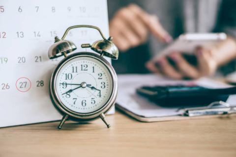 Image of an alarm clock in front of a calendar with a background of a person's hands using a calculator.