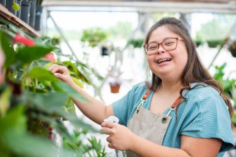 Image of employee with disabilities working at a nursery. She has a spray bottle in her left hand and she is smiling at the camera. 