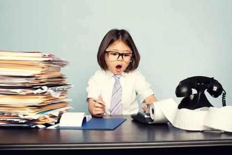 Image of a young student dressed as an accountant and in shock over the results from an adding machine. She has an old rotary phone to one side, and a stack of manilla folders and papers to the other.