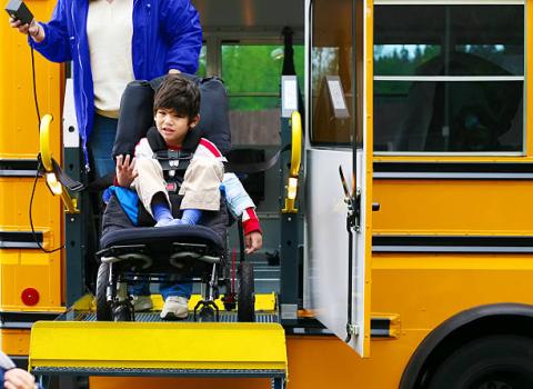 Image of a student in a wheelchair who is being lowered by a lift from a school bus.