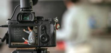 Image of video camera in the forefront pointing to a speaker or instructor in the background. The speaker's image is blurred.